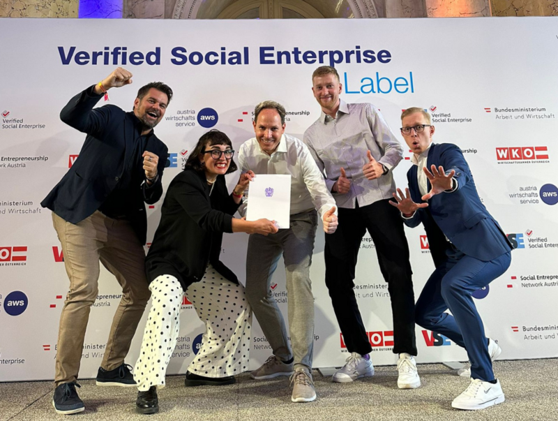 Wolfgang Kowatsch, Fides Raffel, Michael Aumann, Sebastian Stoll und Michael Zakall posieren freudig mit dem Label/Zertifikat "Verified Social Enterprise" vor einer Logowand. Logos WKO, SENA, aws, Bundesministerium für Arbeit und Wirtschaft