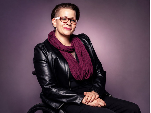 Foto von Claudia Miller mit kurzem braunem Haar, Brille, lila Schal und Lederjacke. Sie sitzt in einem Rollstuhl.