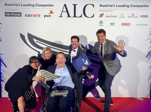 myAbility Mitarbeiter:innen lachen in die Kamera und machen freudige Gesten, den ALC Award in der Hand