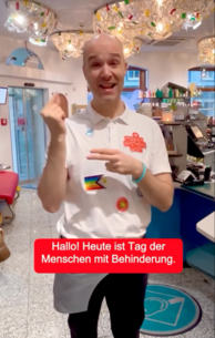Person steht in der Apotheke und kommuniziert mit Gebärden. In den Untertitel steht: "Hallo! Heute ist Tag der Menschen mit Behinderung."