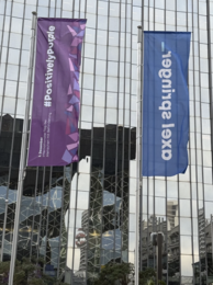 Zwei Fahnenmaste vor einem großen Gebäude. Eine lila Fahne mit "#PositivelyPurple", eine blaue Fahne mit "Axel Springer"