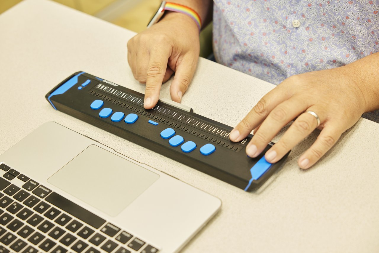 two hands using an electronic Braille display, next to a laptop