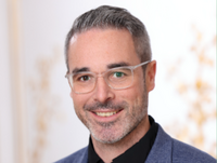 Alt-Text: Porträt eines lächelnden Mannes mit kurzen grauen Haaren und Brille. Er trägt ein schwarzes Hemd und ein blaues Sakko.
